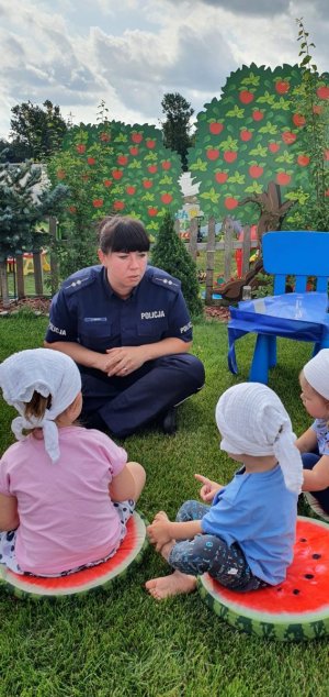 Zdjęcie kolorowe. Policjantka siedząca na trawie rozmawia z dziećmi
