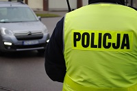 Zdjęcie kolorowe. Na pierwszym planie widać od tyłu policjanta w kamizelce odblaskowej z napisem POLICJA, w oddali srebrny samochód
