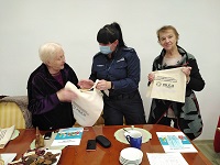 Zdjęcie kolorowe. Policjanta pomiędzy seniorkami pozuje do zdjęcia. Seniorka z lewej strony trzyma torbę z odblaskowym paskiem