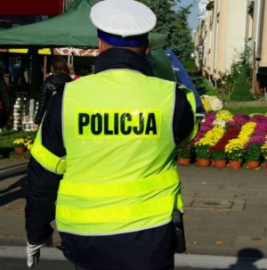 Zdjęcie kolorowe. Policjant stojący placami w kamizelce odblaskowej z napisem policja stoi na tle stoisk z kwiatami i zniczami przed cmentarzem