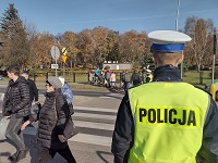 Zdjęcie kolorowe. Policjant obserwuje ruch na przejściu dla pieszych. W tle drugi policjant kieruje ruchem