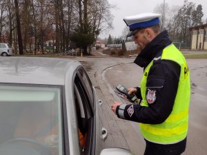Zdjęcie kolorowe. Policjant dokonuje pomiaru trzeźwości kierującego srebrnym pojazdem