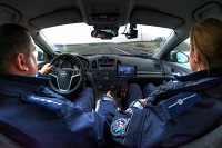 Zdjęcie kolorowe. Zdjęcie wykonane w samochodzie z tylnej kanapy. Na siedzeniu kierowcy i pasażera siedzą policjanci.