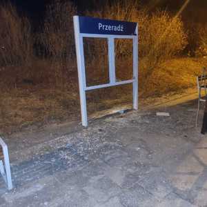 Zdjęcie kolorowe. Zdjęcie zrobione nocą. Na chodniku porozrzucane szkło z tablicy informacyjnej z boku stoi ławka i słup z nazwą miejscowości Przeradź