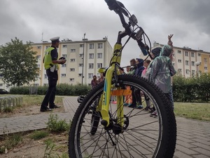 Zdjęcie kolorowe. Na pierwszym planie widoczny jest rower. W tle policjant prowadzący spotkanie z dziećmi.