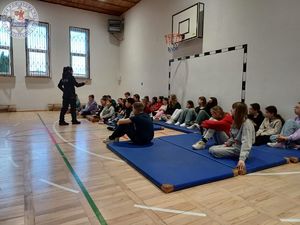 Zdjęcie kolorowe. Policjantka prowadzi zajęcia z uczniami, którzy siedzą na materacach na sali gimnastycznej