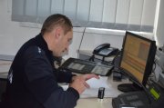 Zdjęcie kolorowe. Policjant siedzący przy biurku - stanowisko dowodzenia. Na biurku stoi włączony komputer, stoi również telefon stacjonarny