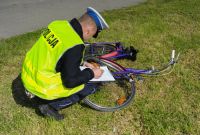 Zdjęcie kolorowe. Umundurowany policjant ruchu drogowego pochylony jest nad rowerem i sporządza dokumentację.